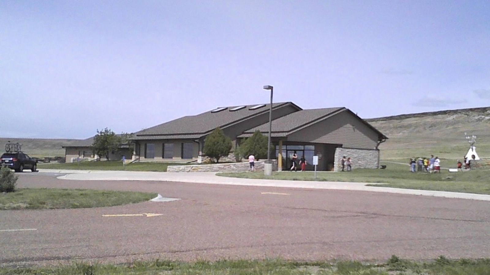 A parking lot sits in front of a grey building as visitors walk around