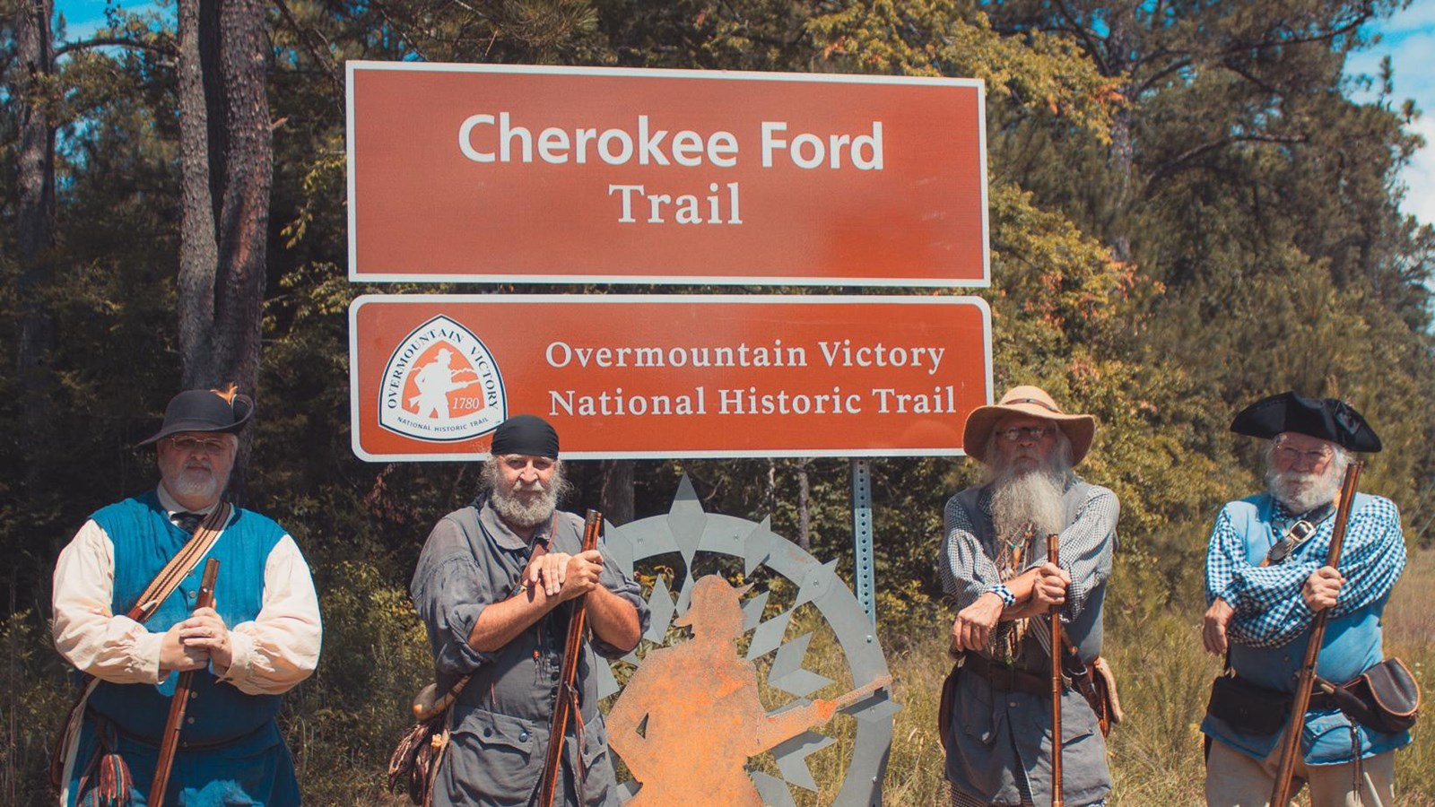 Four men in 1780s frontier costumes stand beside a large brown metal sign