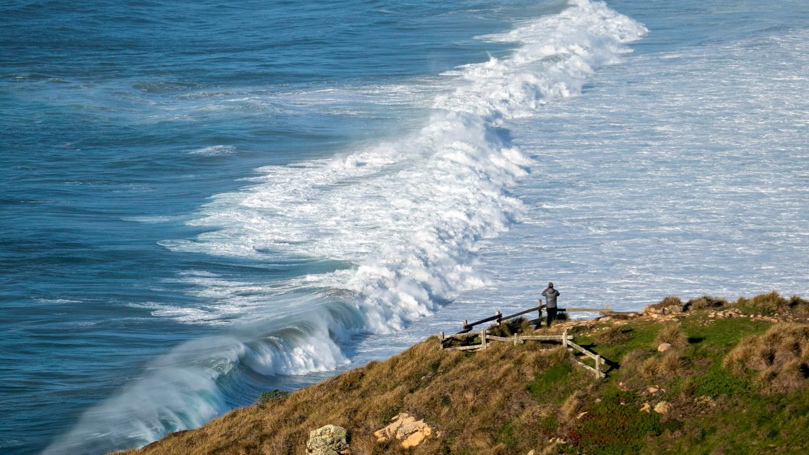 A visitor watches the crashing waves below from a high and fenced-in overlook. 