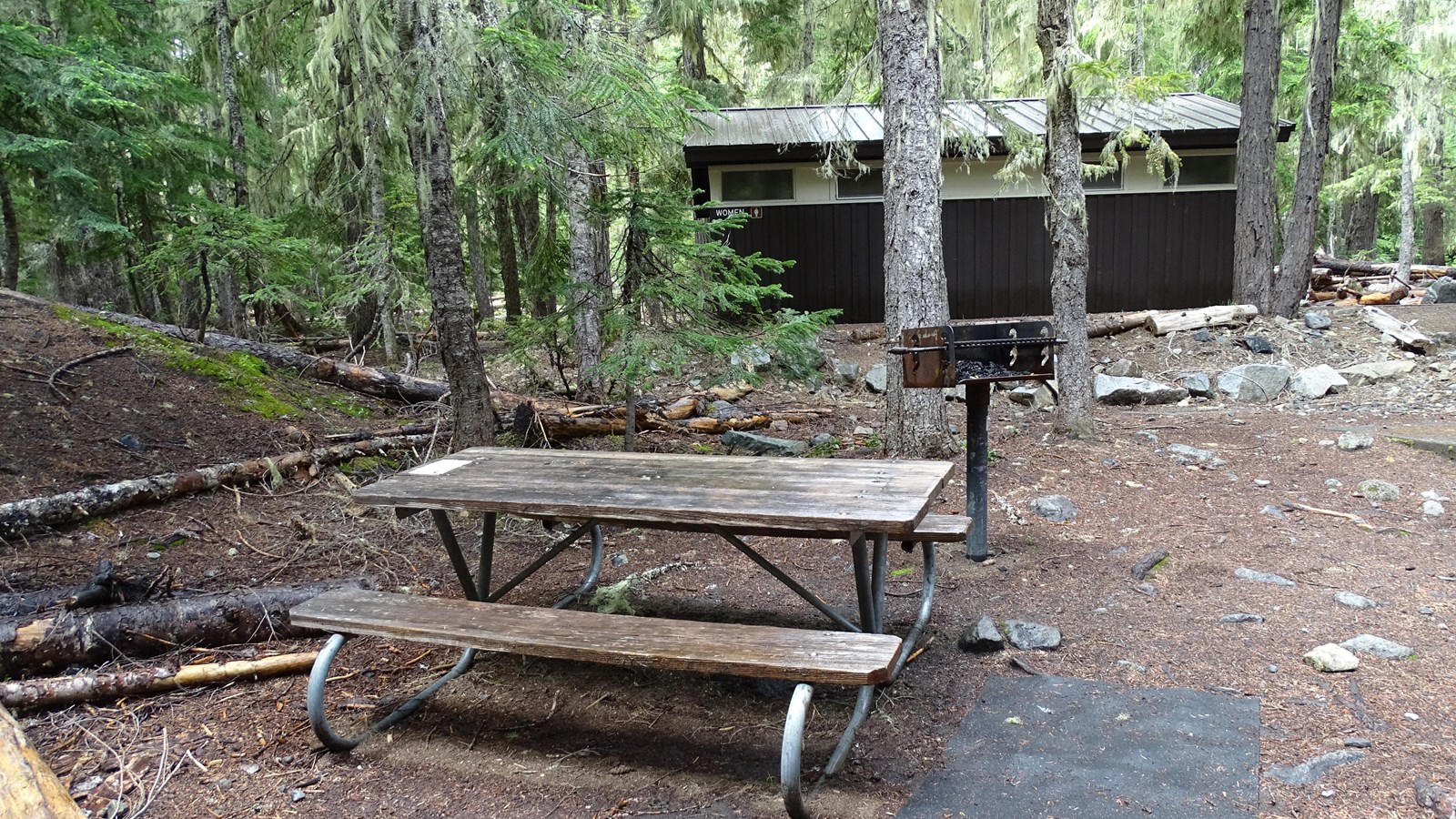 A wooden picnic table with a grill surrounded by trees, and a brown building in the background.