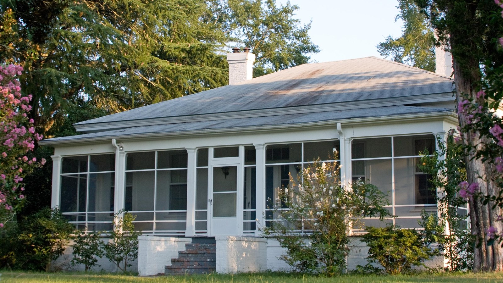 A square white house with trees and flowering shrubs on either side of it.