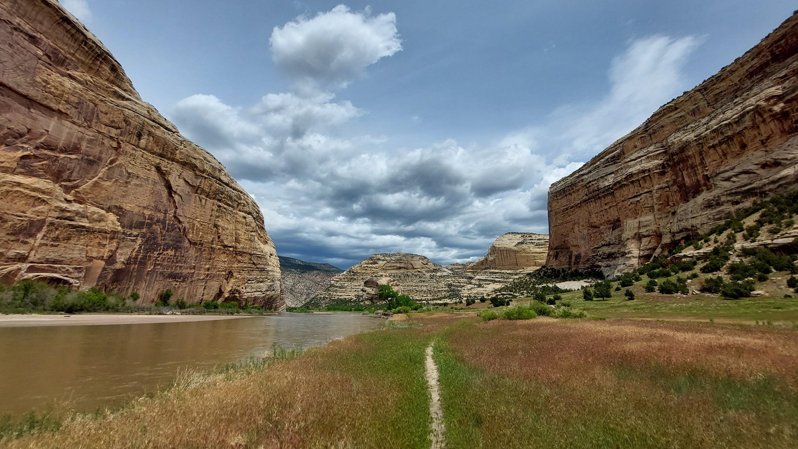 An even-terrain, sandy trail traveling along a river flowing through a steep canyon with sandstone c