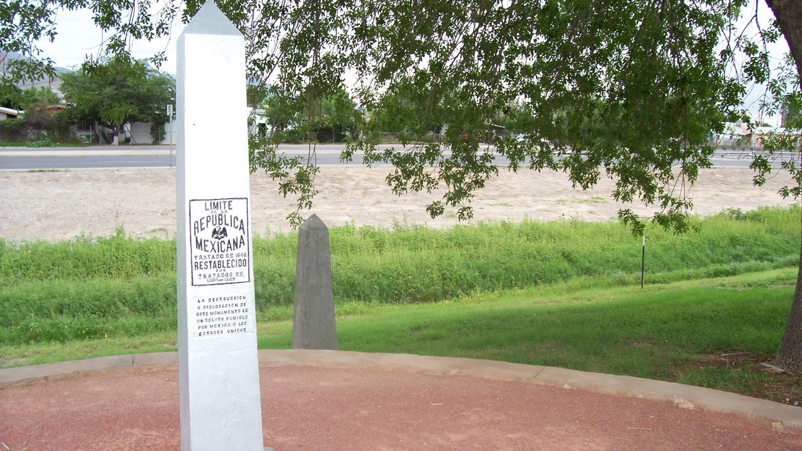Silver colored metal obelisk bearing plaques with text, bolted to ground beside trail