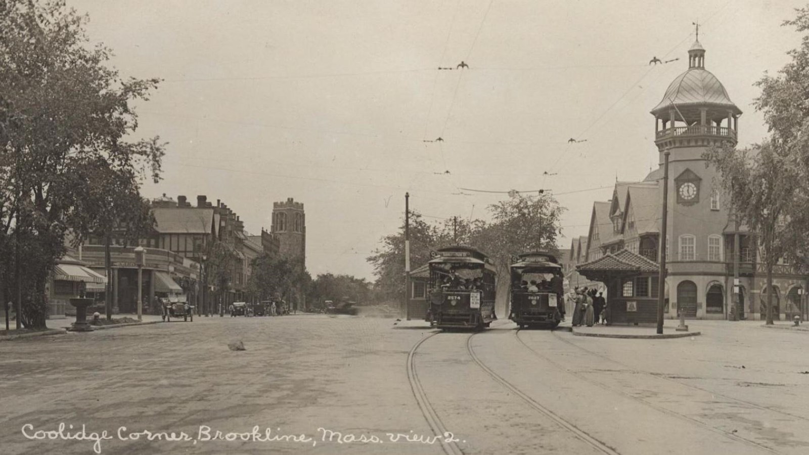 Black and white photograph of wide street, with two trolleys stopped at platforms in the street.