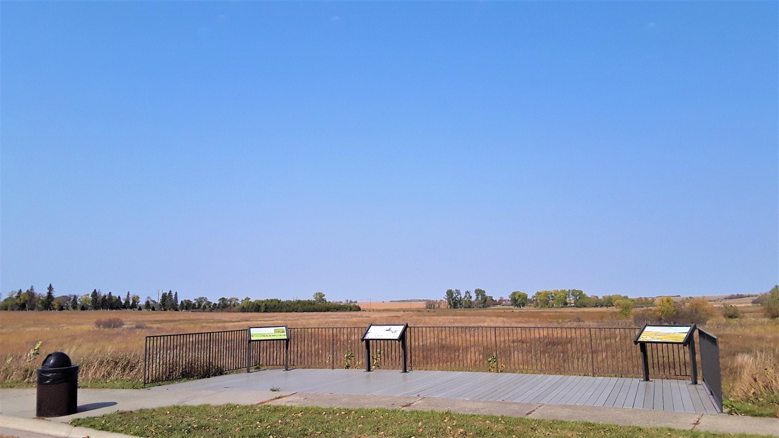 A platform near a curb with 3 waysides and a trash can next to it overlooking prairie grass 
