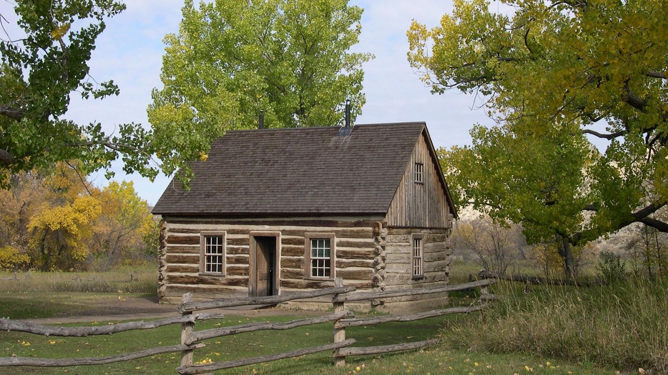 A log cabin sits in a grassy area on the other side of a wooden fence, trees and blue sky behind it.