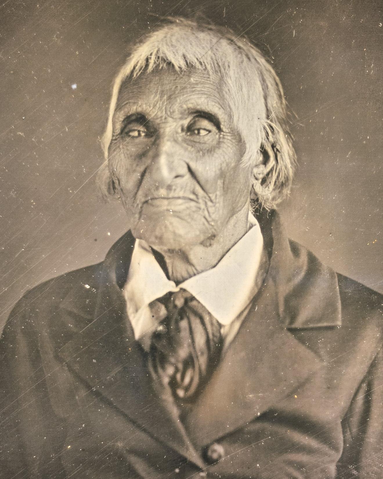 An old man in a black & white portrait. His hair is cropped and white, face worn with heavy lines. 