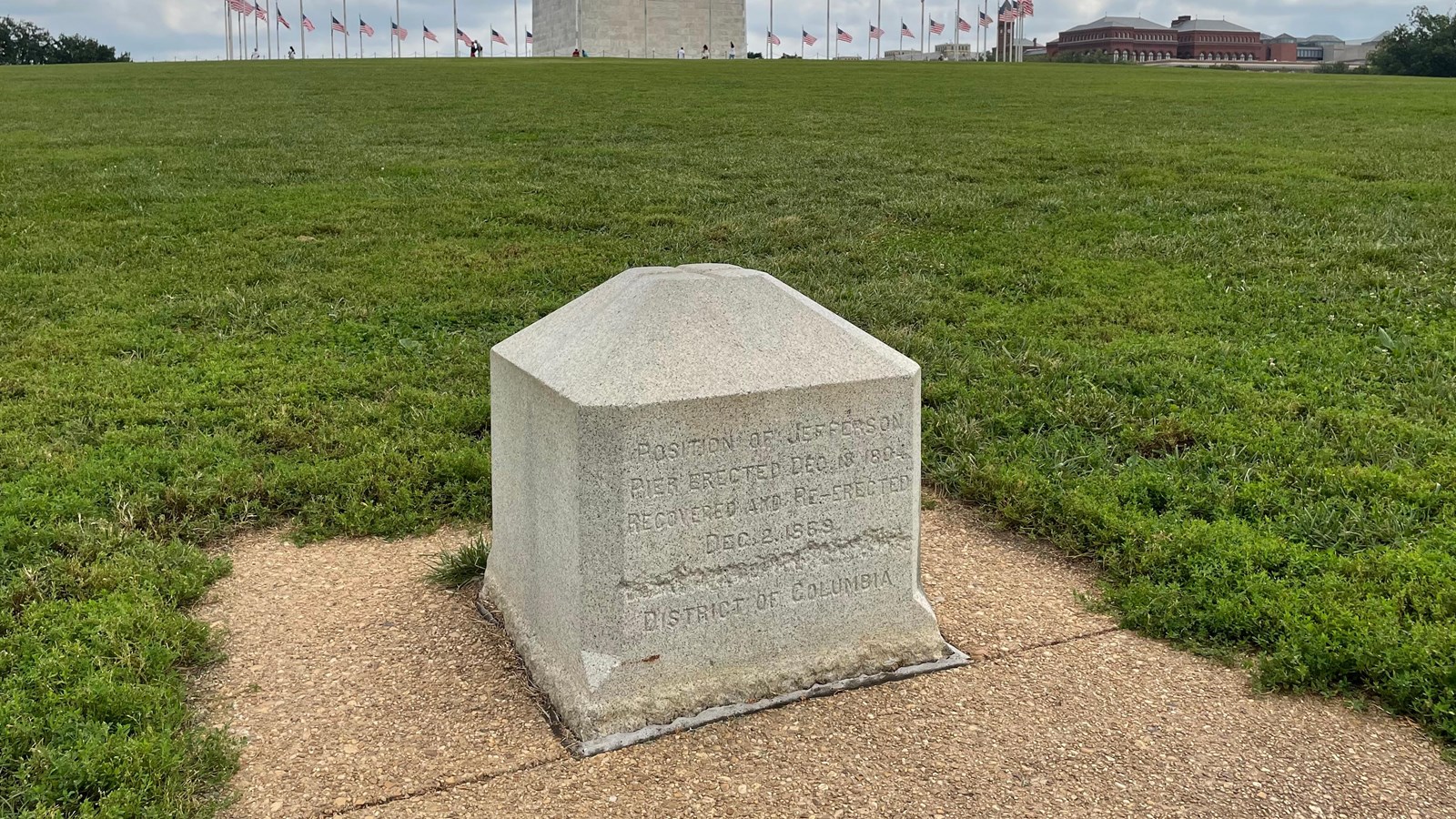 A stone block with words carved into the face on a concrete pad, surrounded by green grass.
