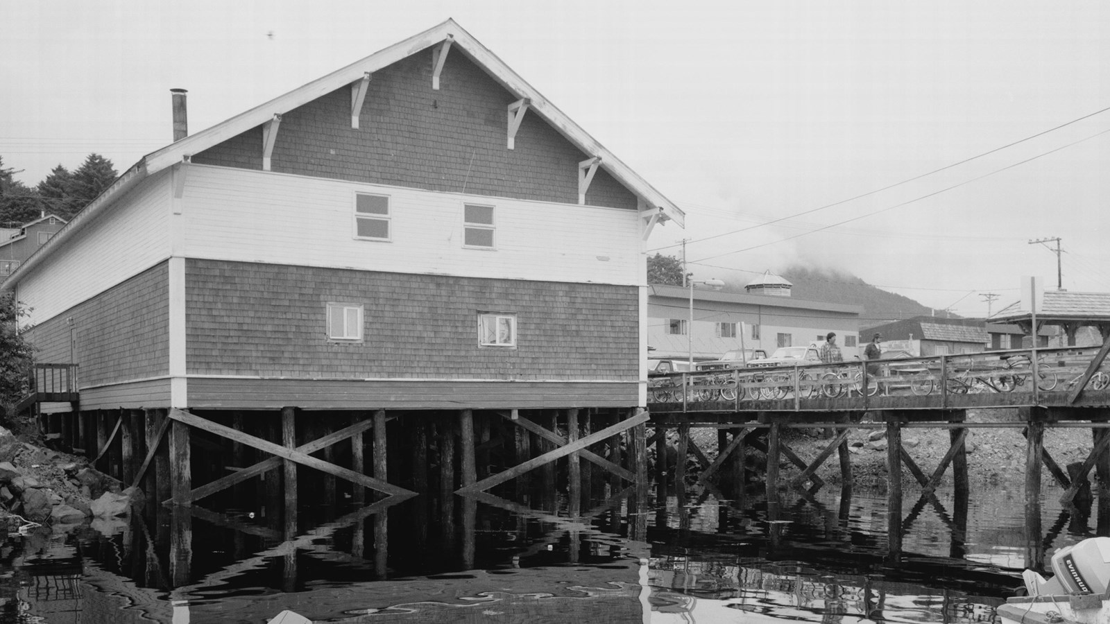 Black and white photo of a two story building on wood pilings over water.