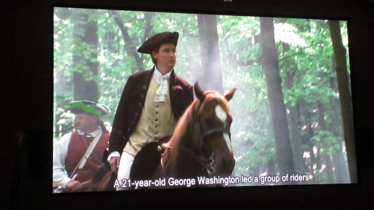 Screen shot of a movie showing a young colonial officer on a horse followed by a man in ar red vest