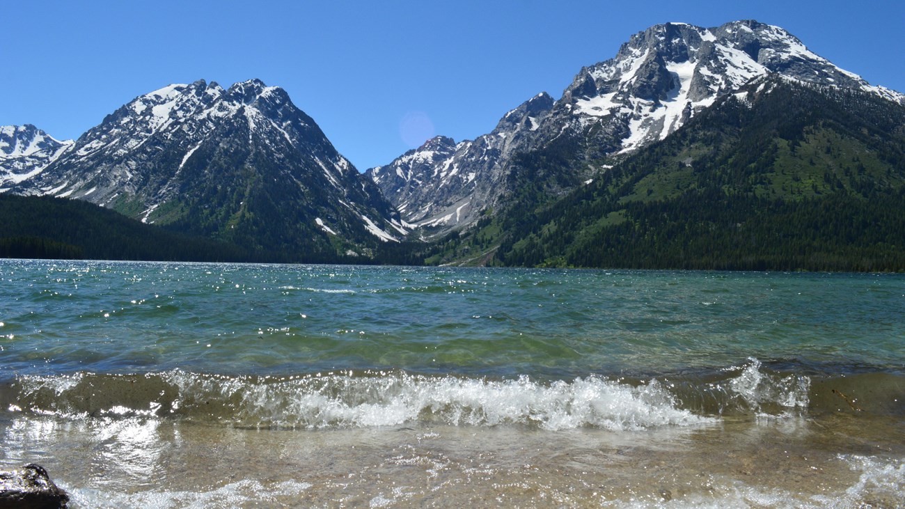 Waves crash on the shore of a clear, blue lake at the base of a mountain range.