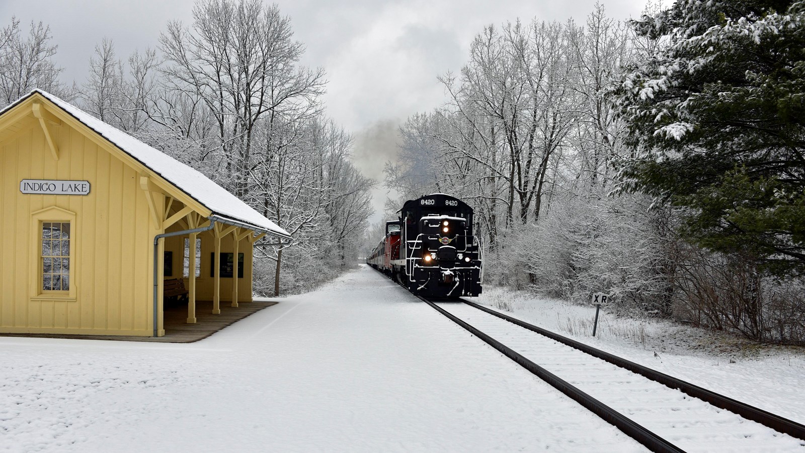 Snow covers trees and yellow station. At right, dark historic train disappears from view on tracks.