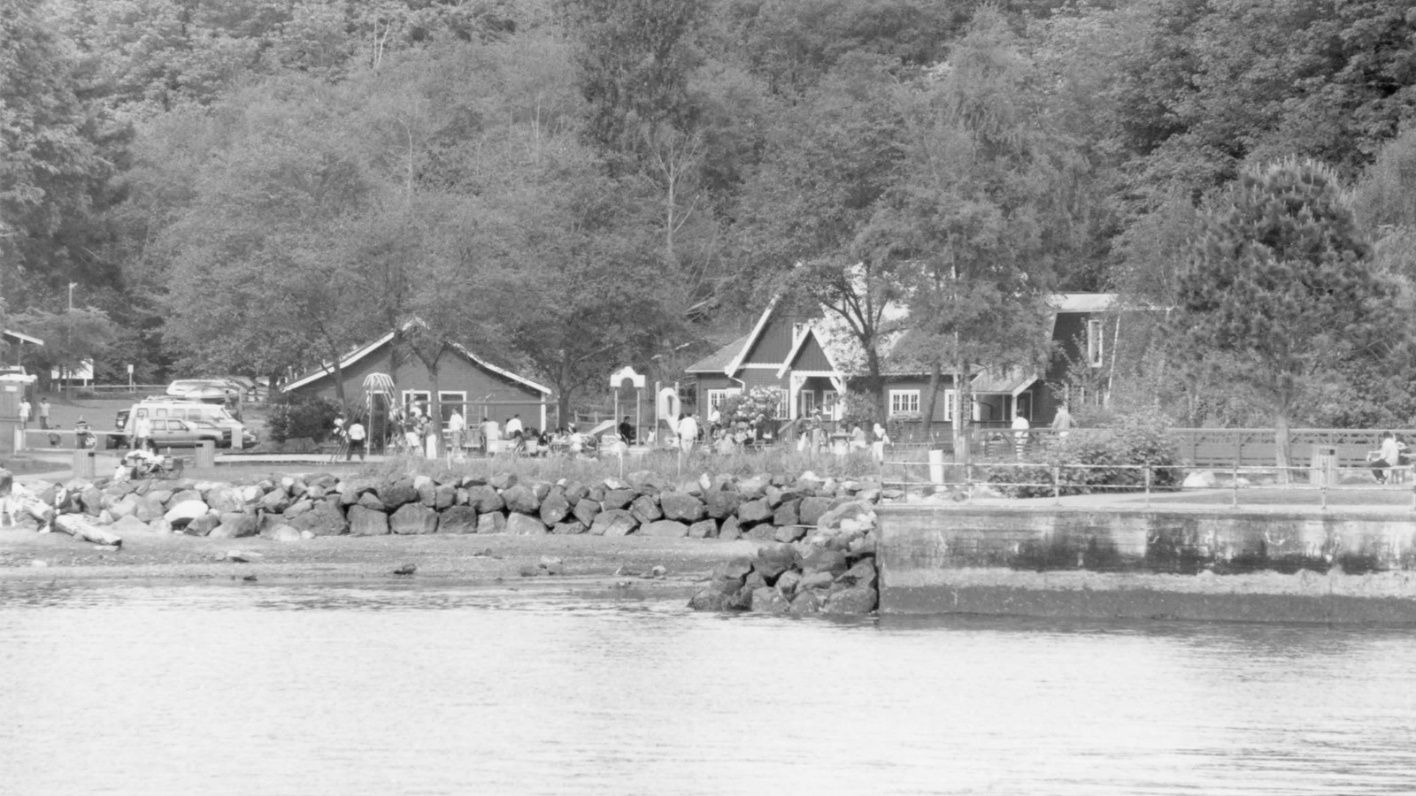 Rocky beach with barn-style buildings with white trim, pedestrians, and parked cars on the left
