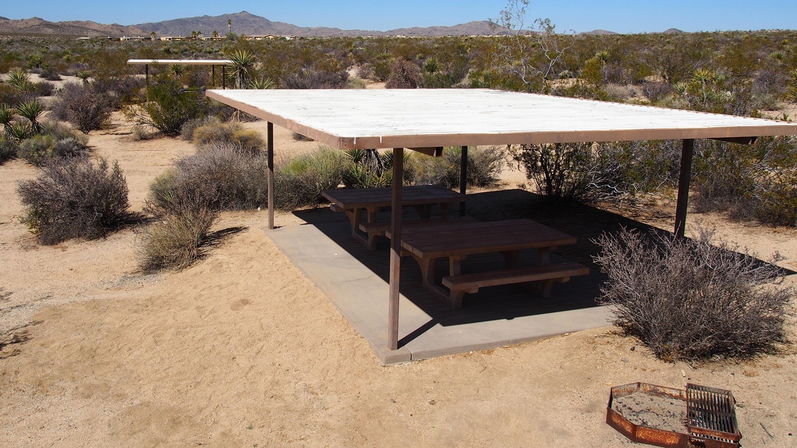 A covered picnic area with two picnic tables and a grill surrounded by desert shrubs.