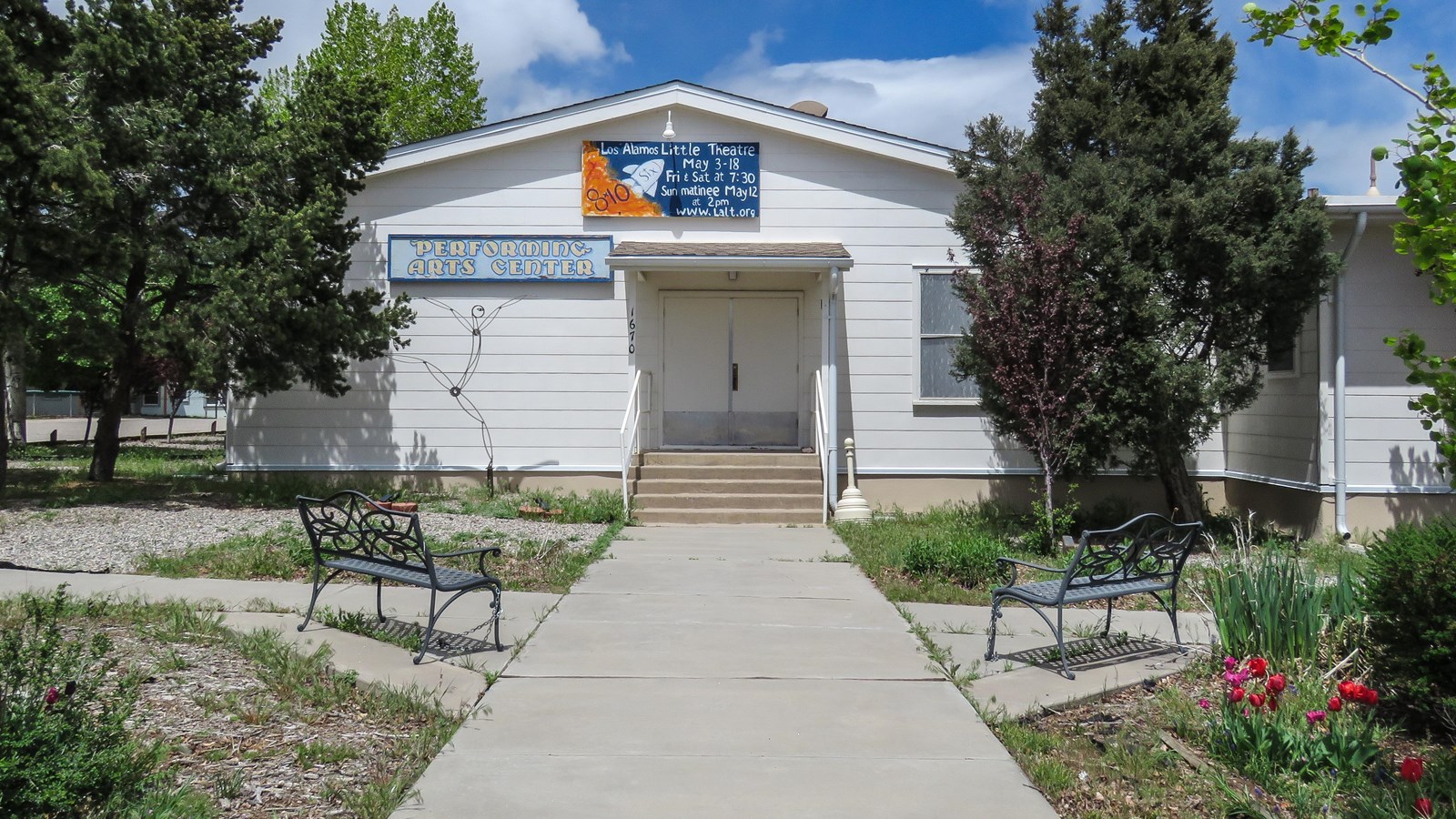 At the end of a wide sidewalk, a white sided building with double doors sits under a cloudy sky.