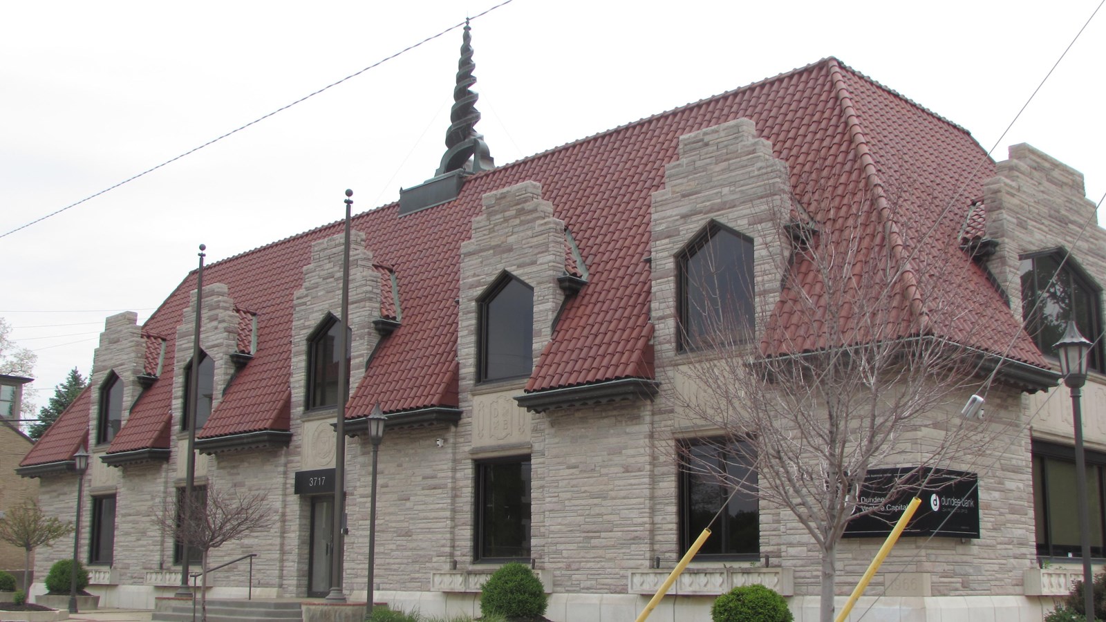 Steeply pitched roof with series of dormers interrupting red tile roof. Stone façade
