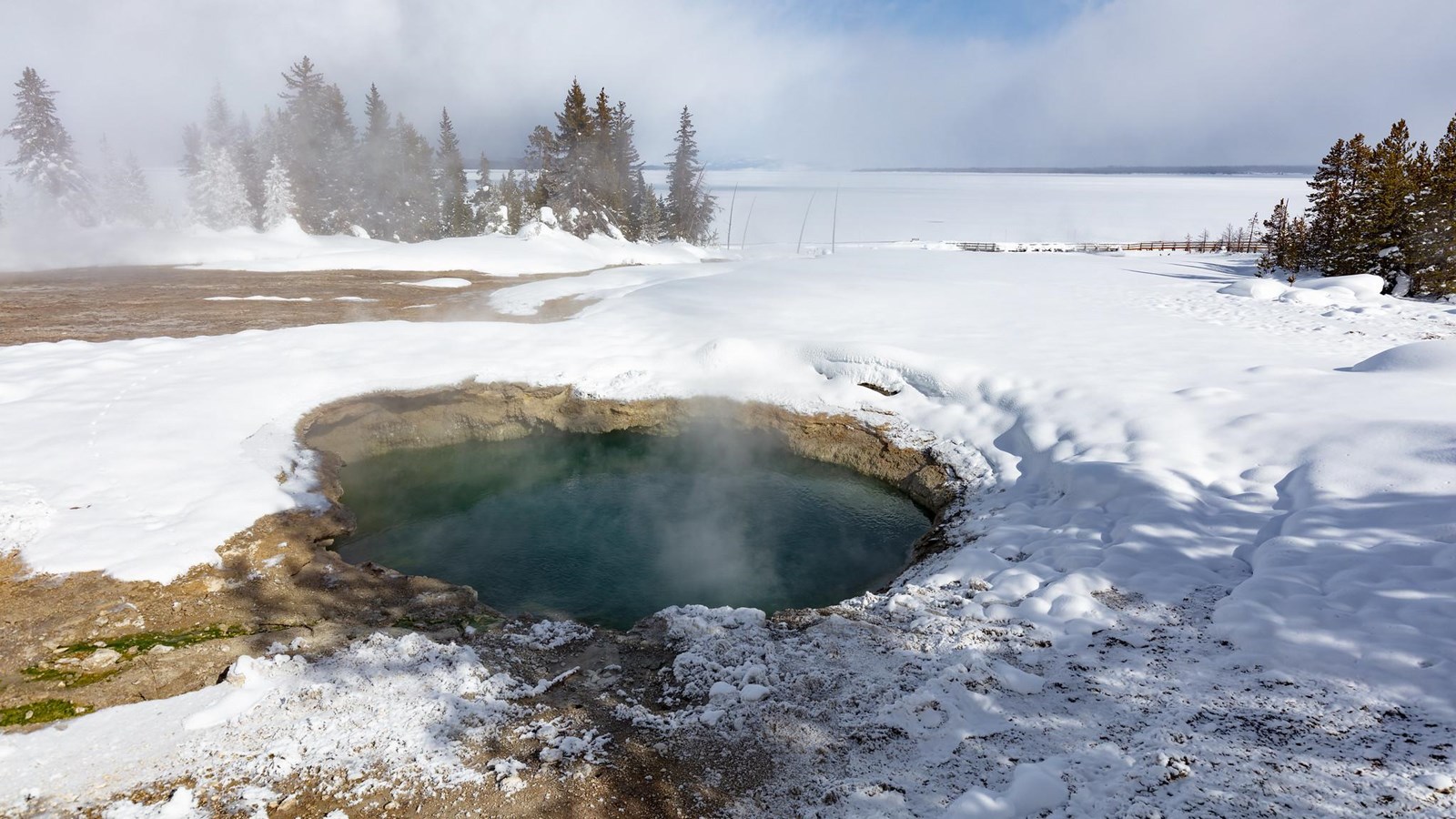 Steam rises from a hot spring in a geyser basin covered in snow along the shore of a frozen lake.