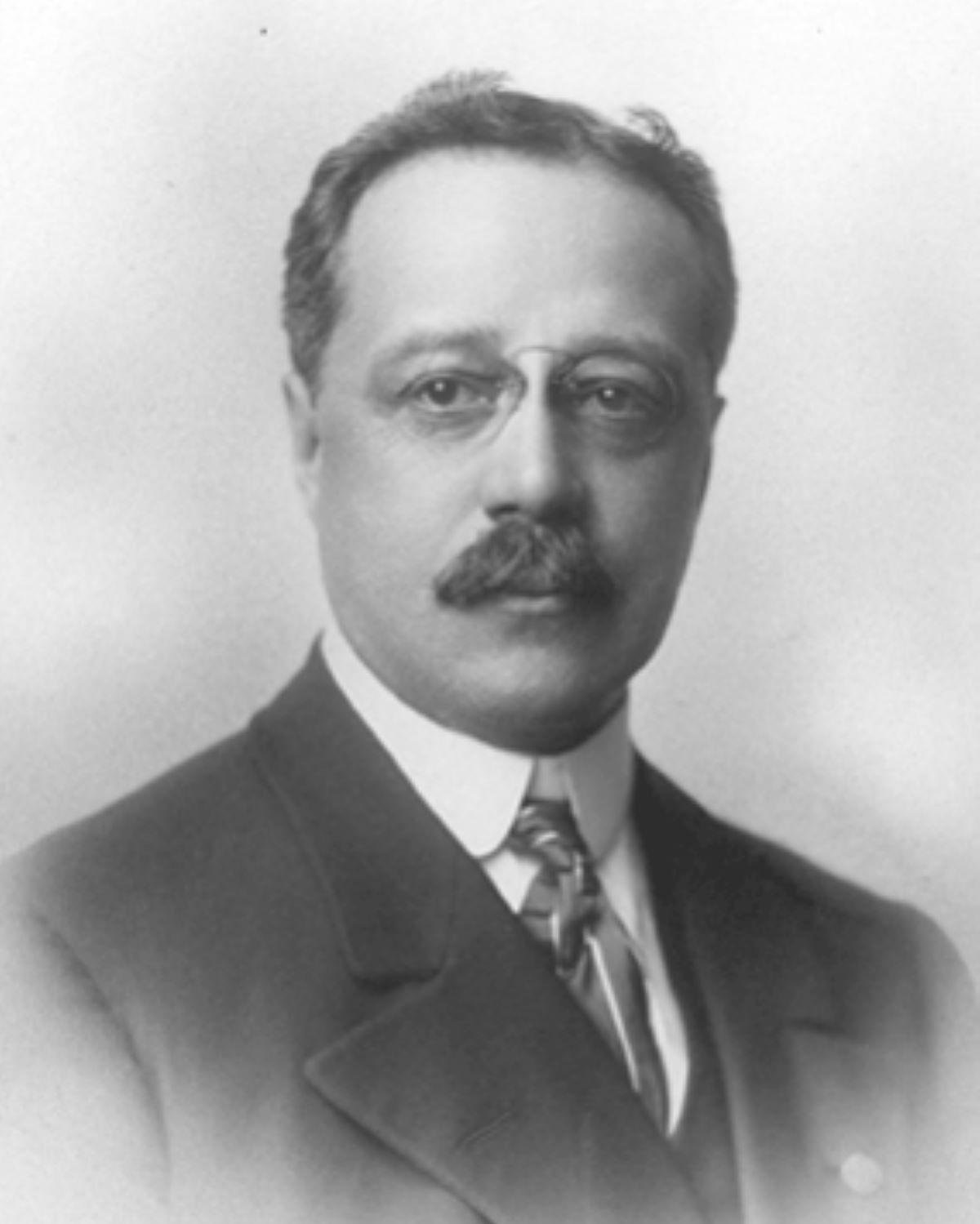 Black and white headshot of a man with a mustache, wearing a suit, tie, and glasses. 