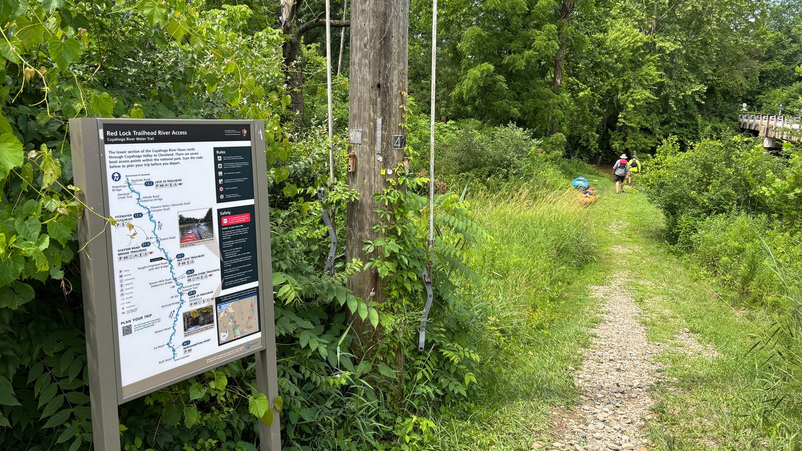 Informational sign to left of a mown grass path; two people in life vests walk along it past kayaks.