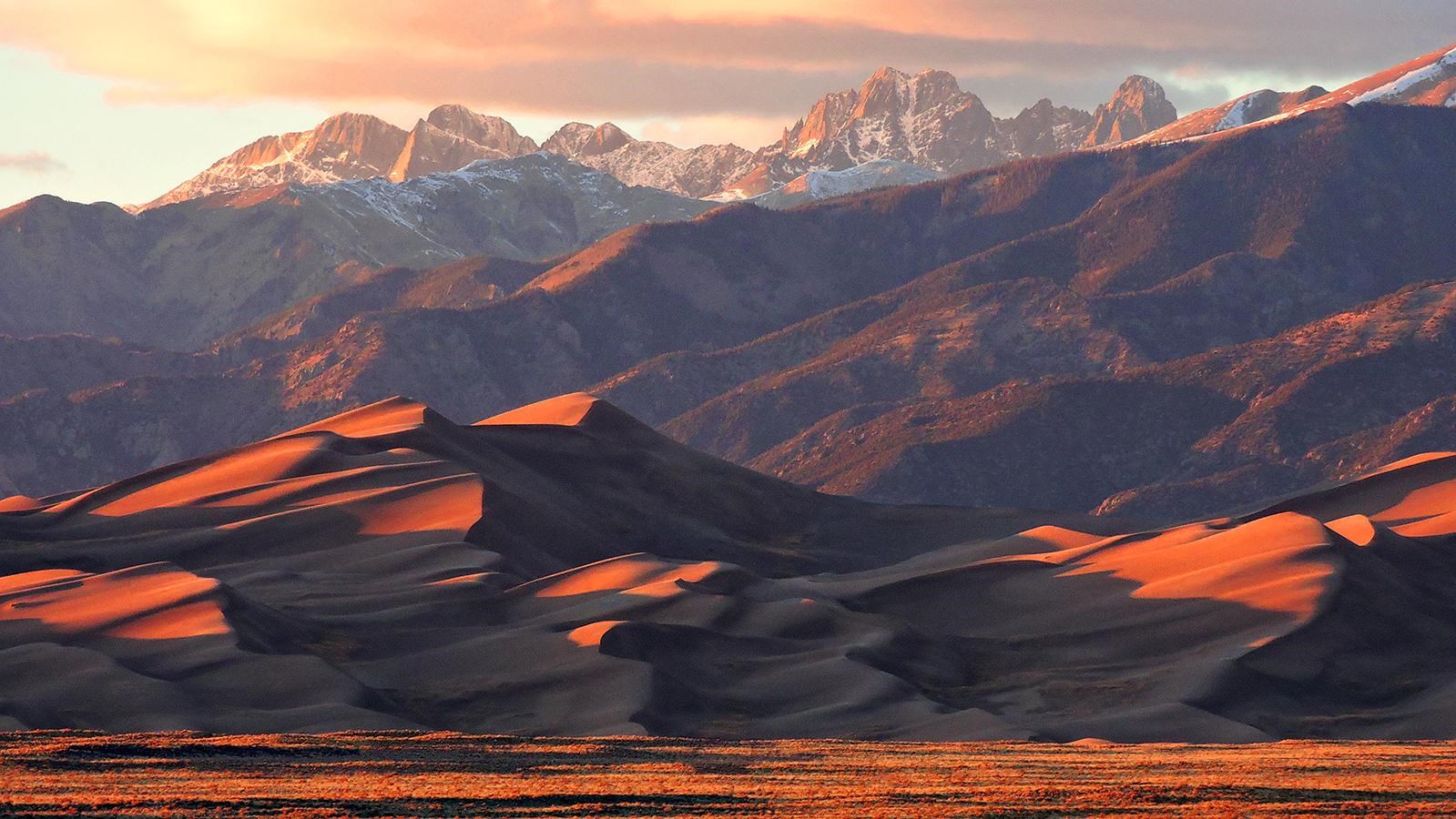 A huge pyramid-shaped dune and jagged, snow-capped peaks at sunset