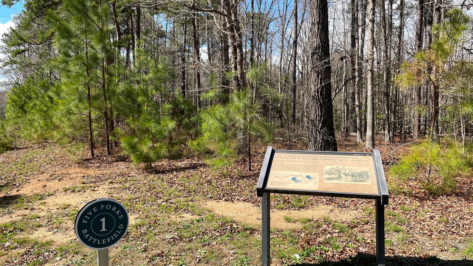 Wayside and smaller round sign in front of a wooded area.