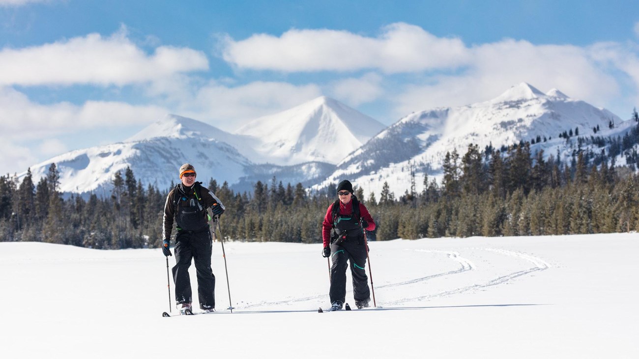Two skiers travel across a flat meadow with mountains in the distance.