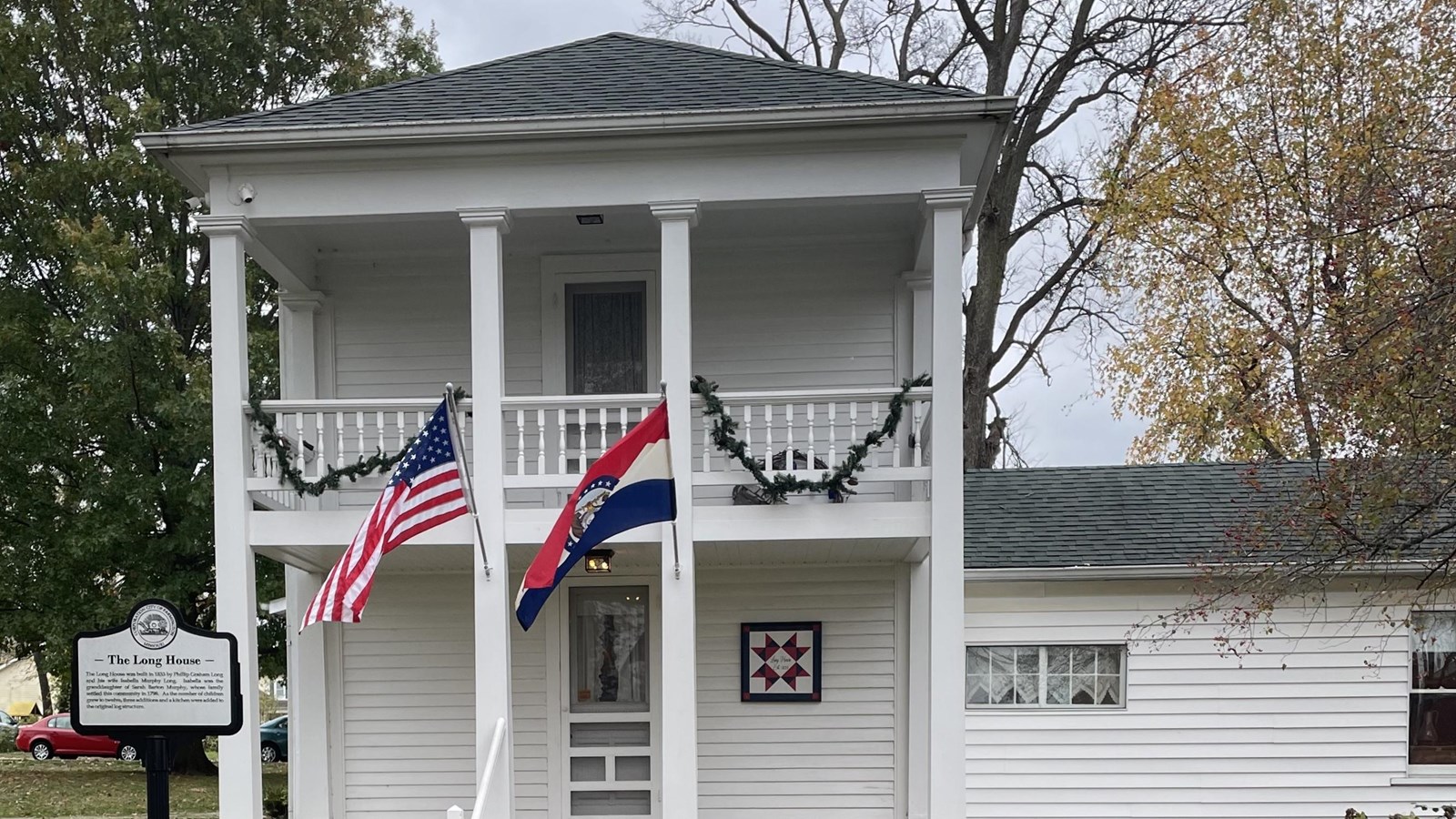 The front facade of a historic, two-story colonial home with banners hanging from the top veranda.