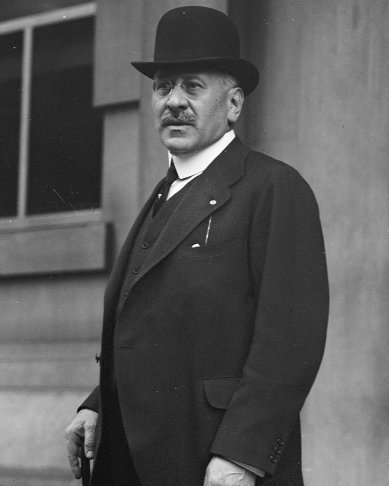 A man in a black suit stands in front of a wooden building in a black and white photograph.