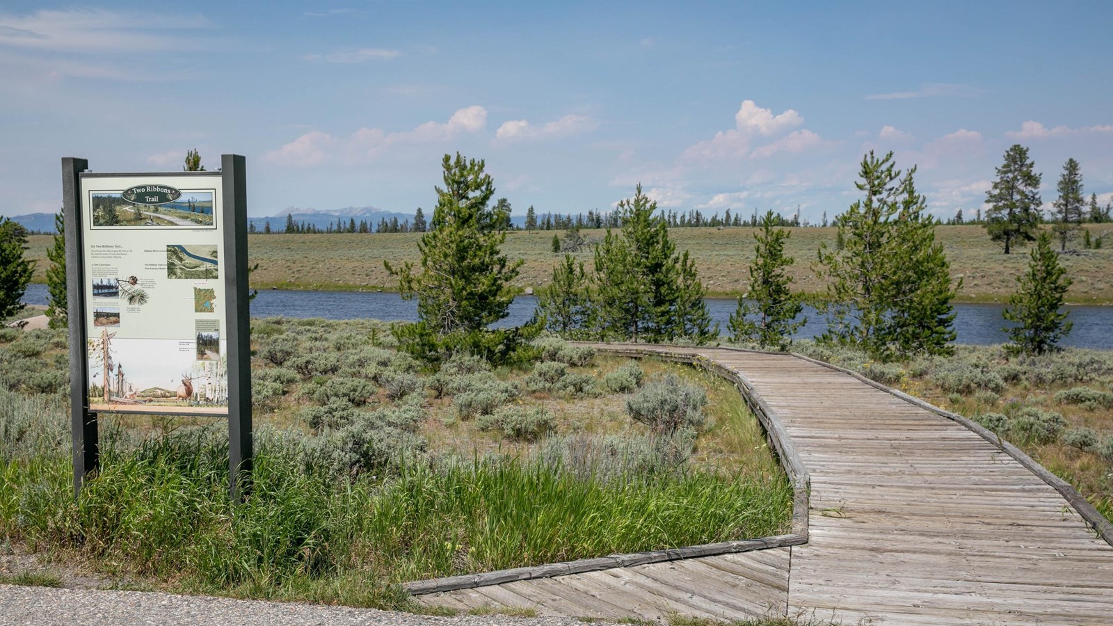 A boardwalk travels through the sagebrush towards a river with mountains in the distance.