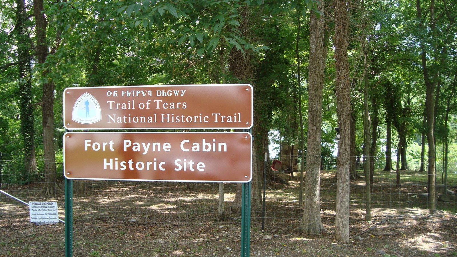 A large brown sign sits in front of a wooded area enclosed by a chain link fence.
