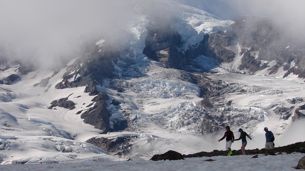 Three hikers crossing a large patch of snow with Mount Rainier in the background.
