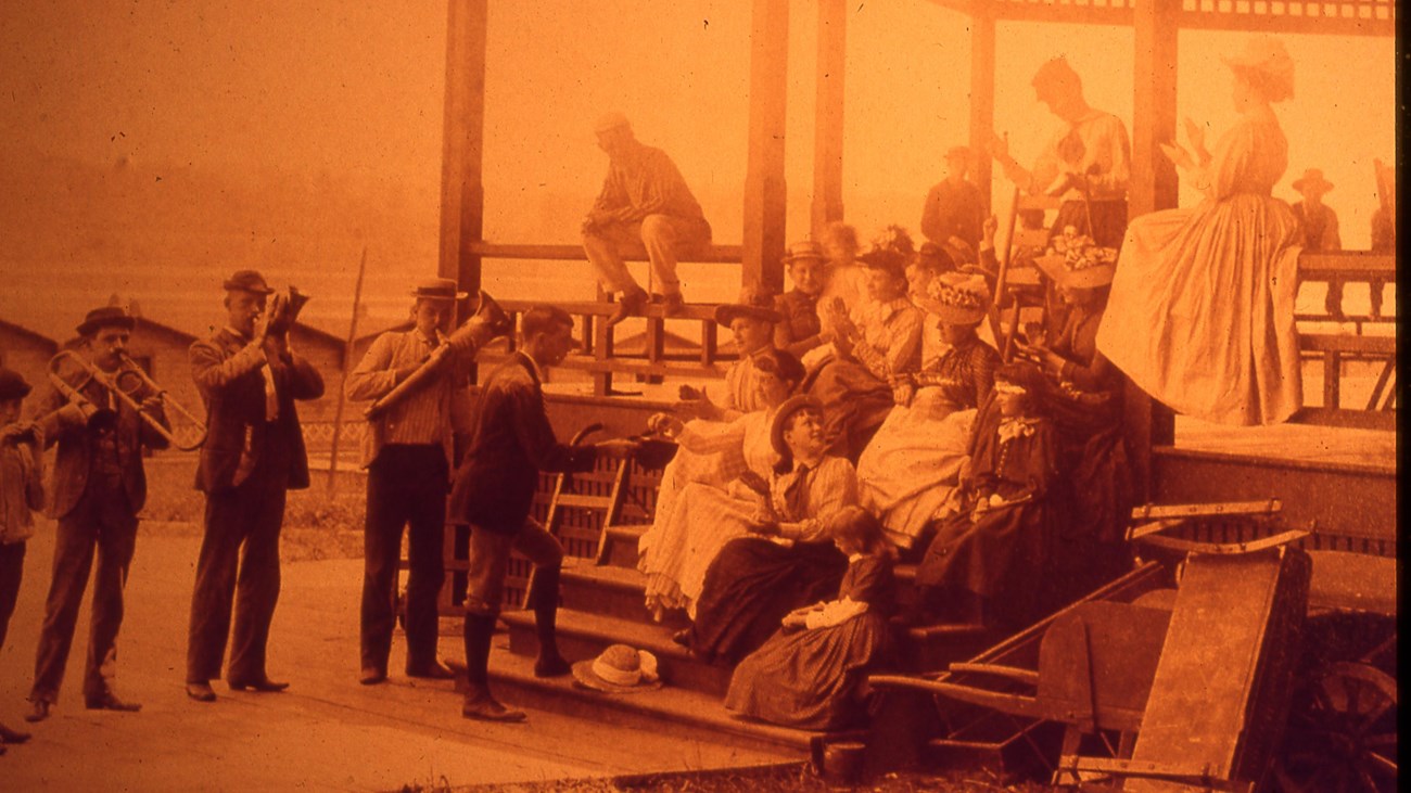 A group of people in period clothing listening to a band play with the lake in the background.