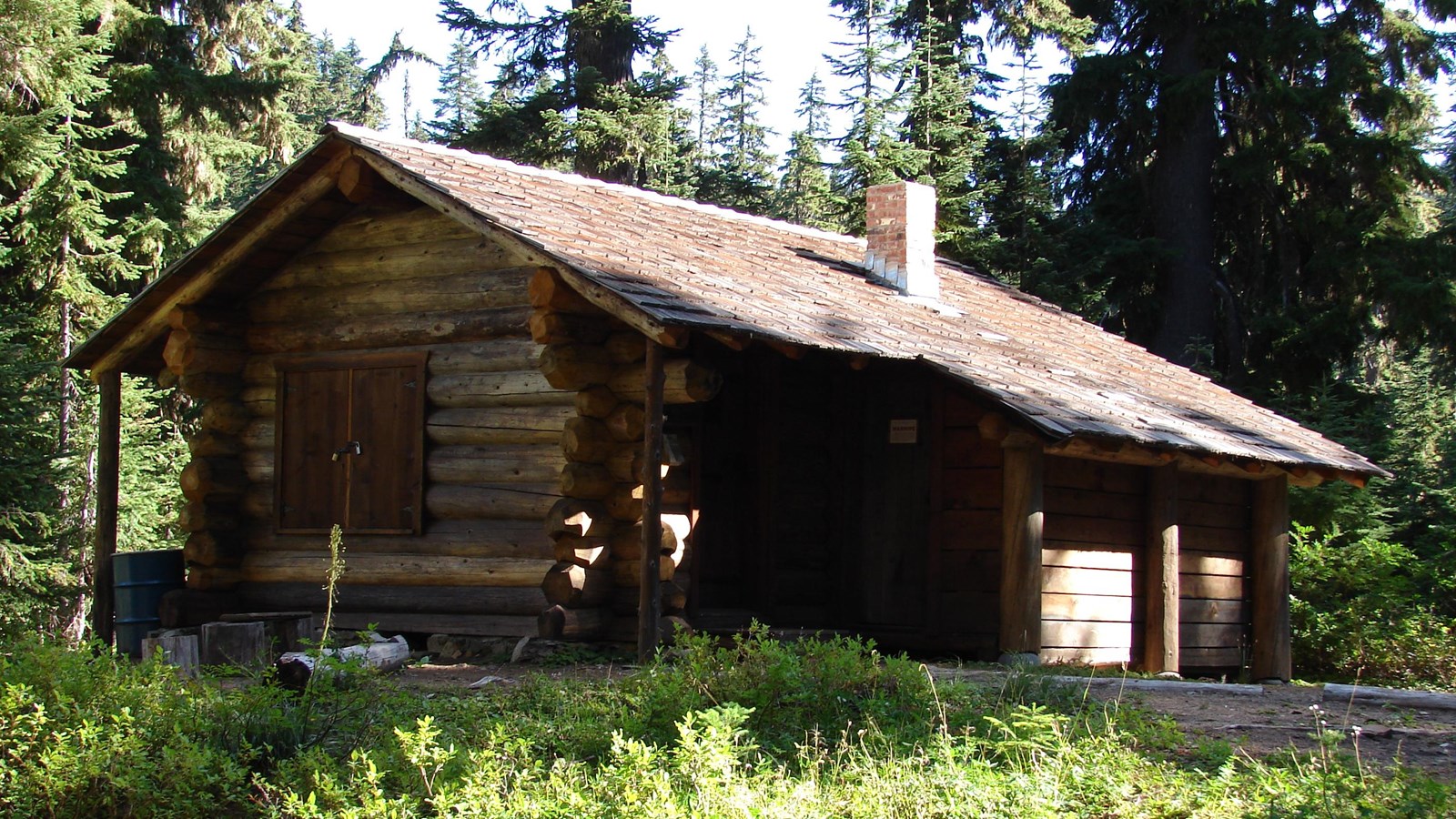 Brown one-story, one-room rectangular log-frame cabin surrounded by trees.