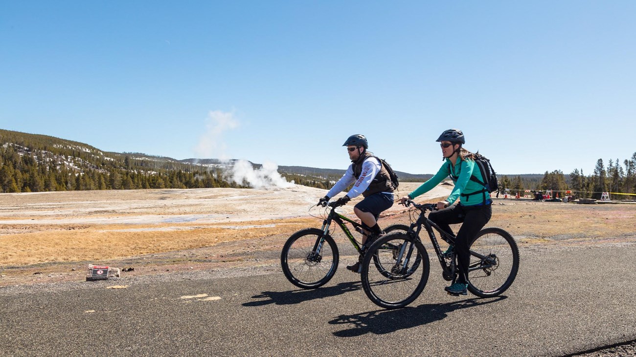 Two people bike on a paved trail past a geyser in a thermal basin.