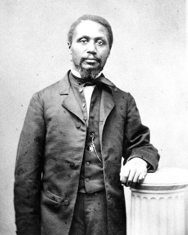 African American man standing next to a column, resting his left arm on the the column. 