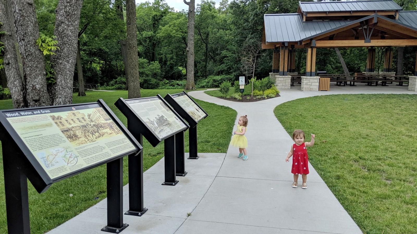A series of exhibit signs line a sidewalk, with two toddles exploring, between a grassy expanse.