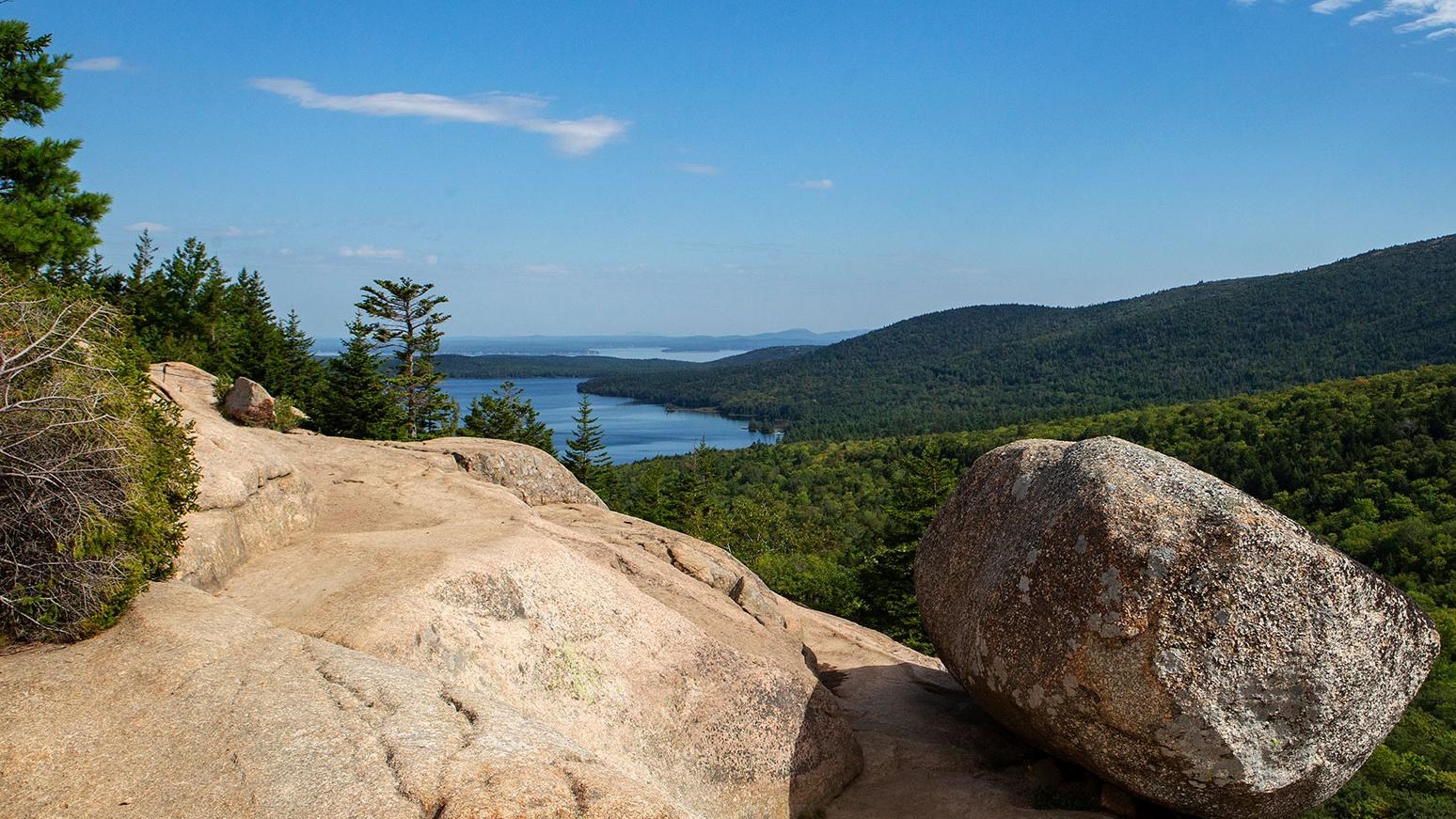 a large round glacial erratic boulder clings to the side of a mountain above woods and waters beyond