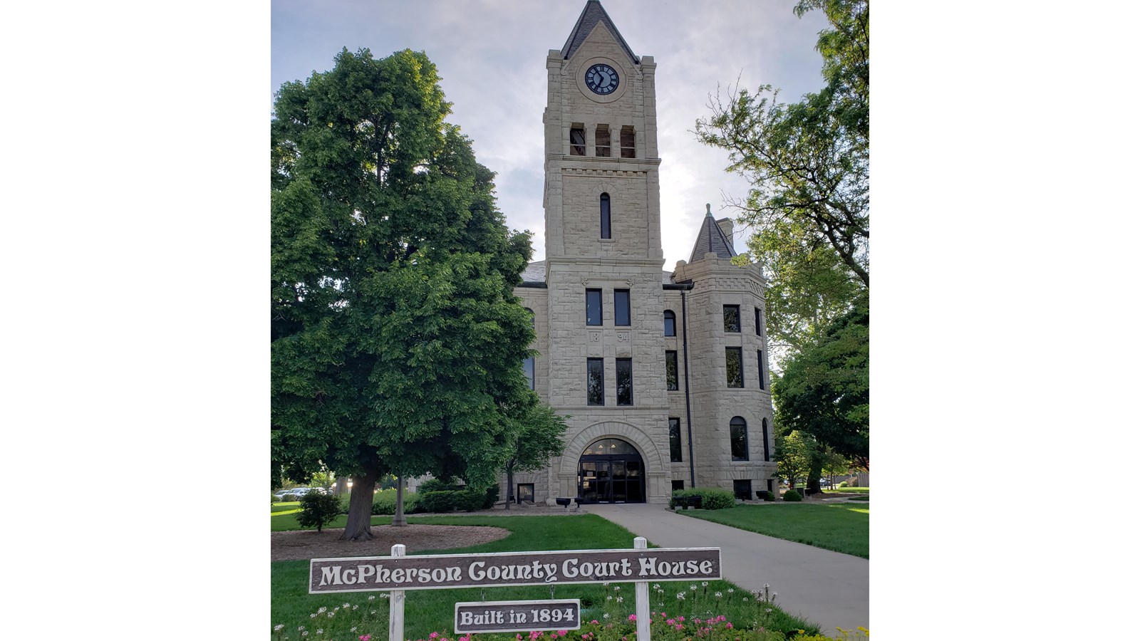 Large stone building with clock tower and a path leading up to it on a sunny day. Green trees