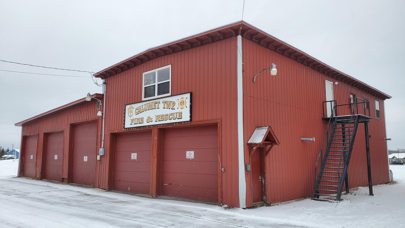 Winter scene of two-story red metal building with one window on top and 5 garage doors.
