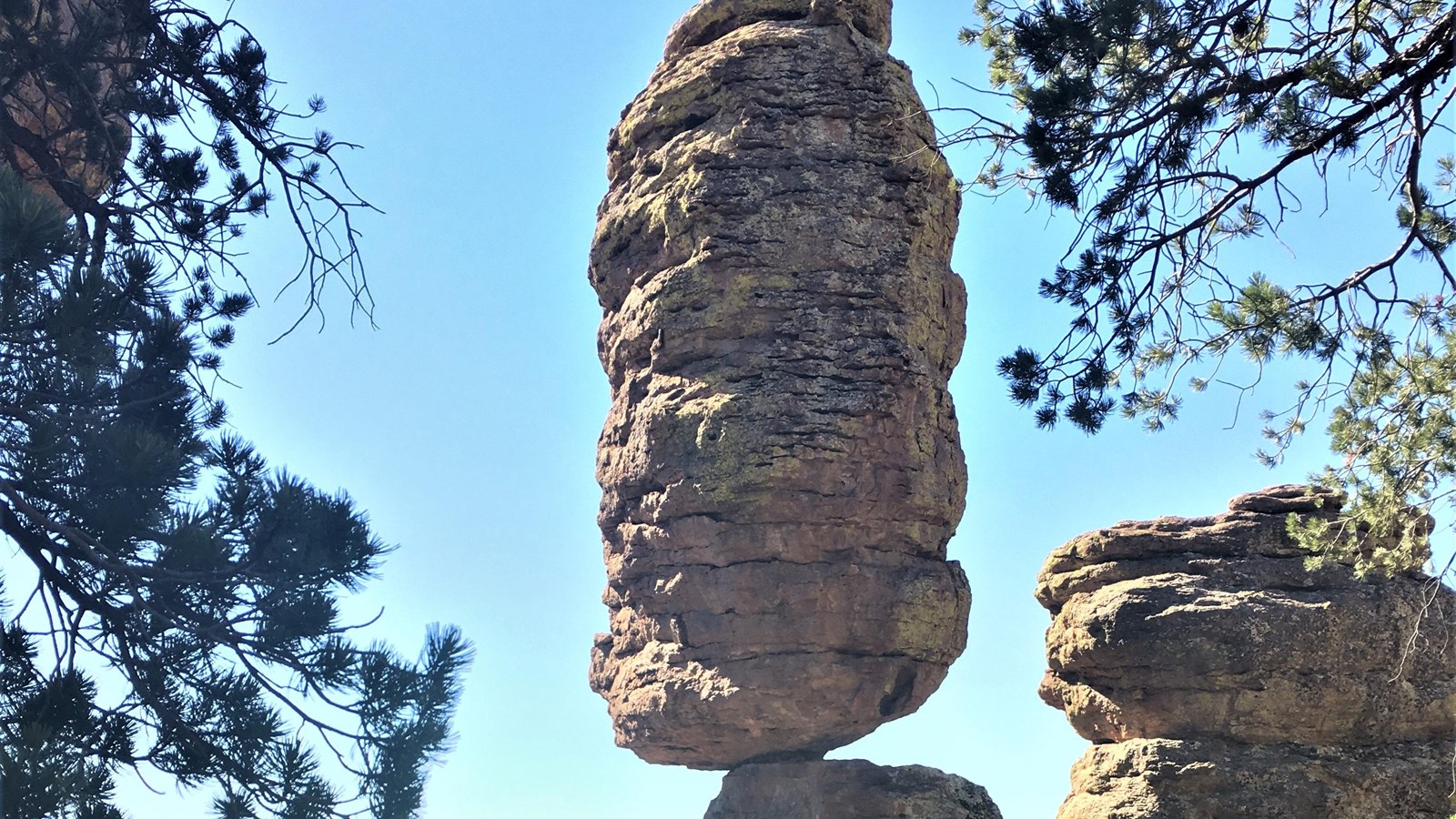 Tall rock balanced on a small point, surrounded by trees and a blue sky