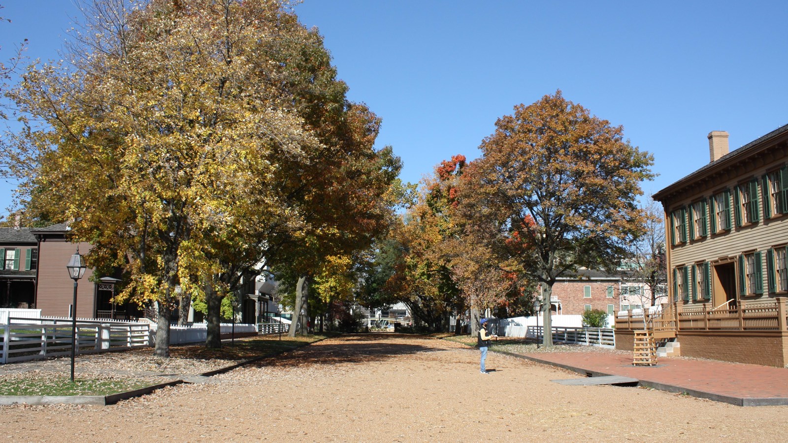 Gravel-street lined with street and wooden boardwalk. The Lincoln Home, tan 2-story house, on right