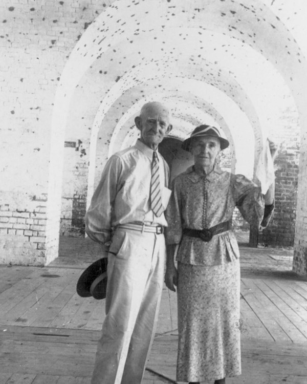 An older woman and an older man stand inside of a brick building with wooden floors.