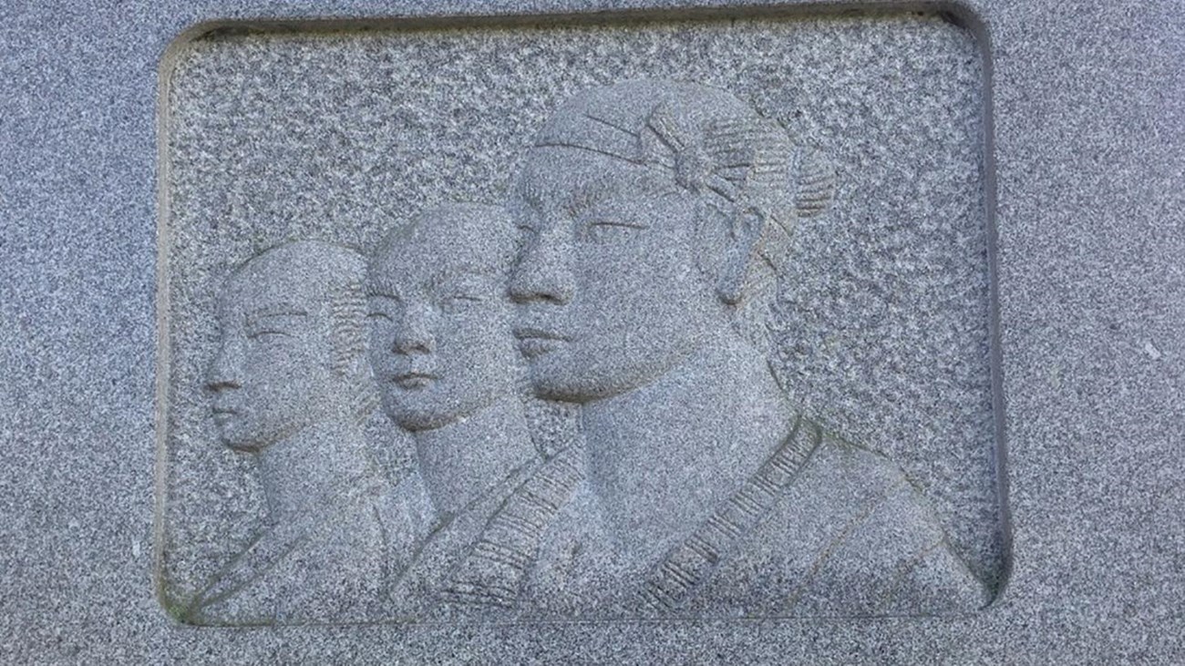 A photo of a stone monument with a carving of three Japanese sailors and text in Japanese.