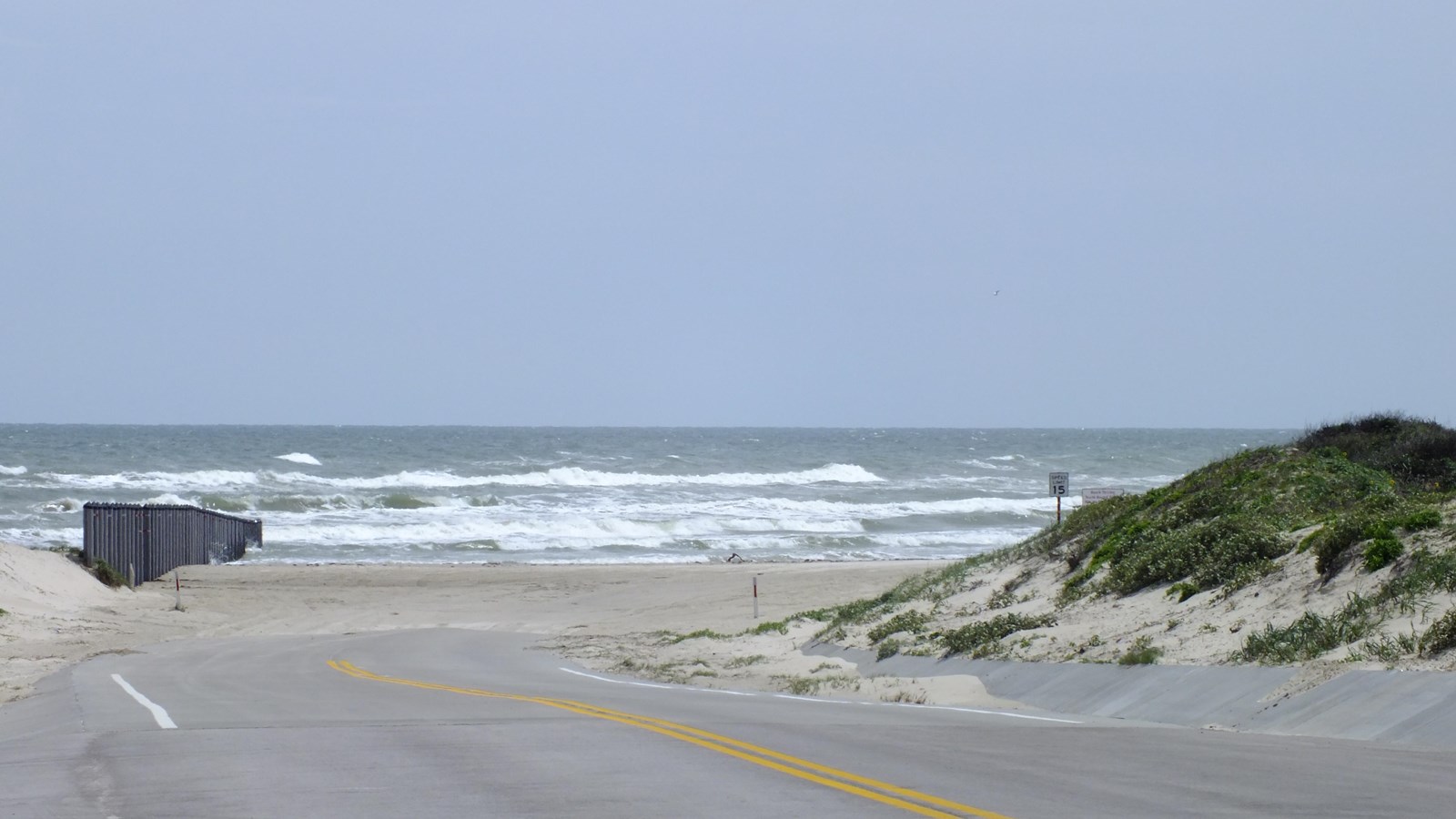 The paved road ends and turns into beach. The gulf is ahead. Bollards block the beach to the left.
