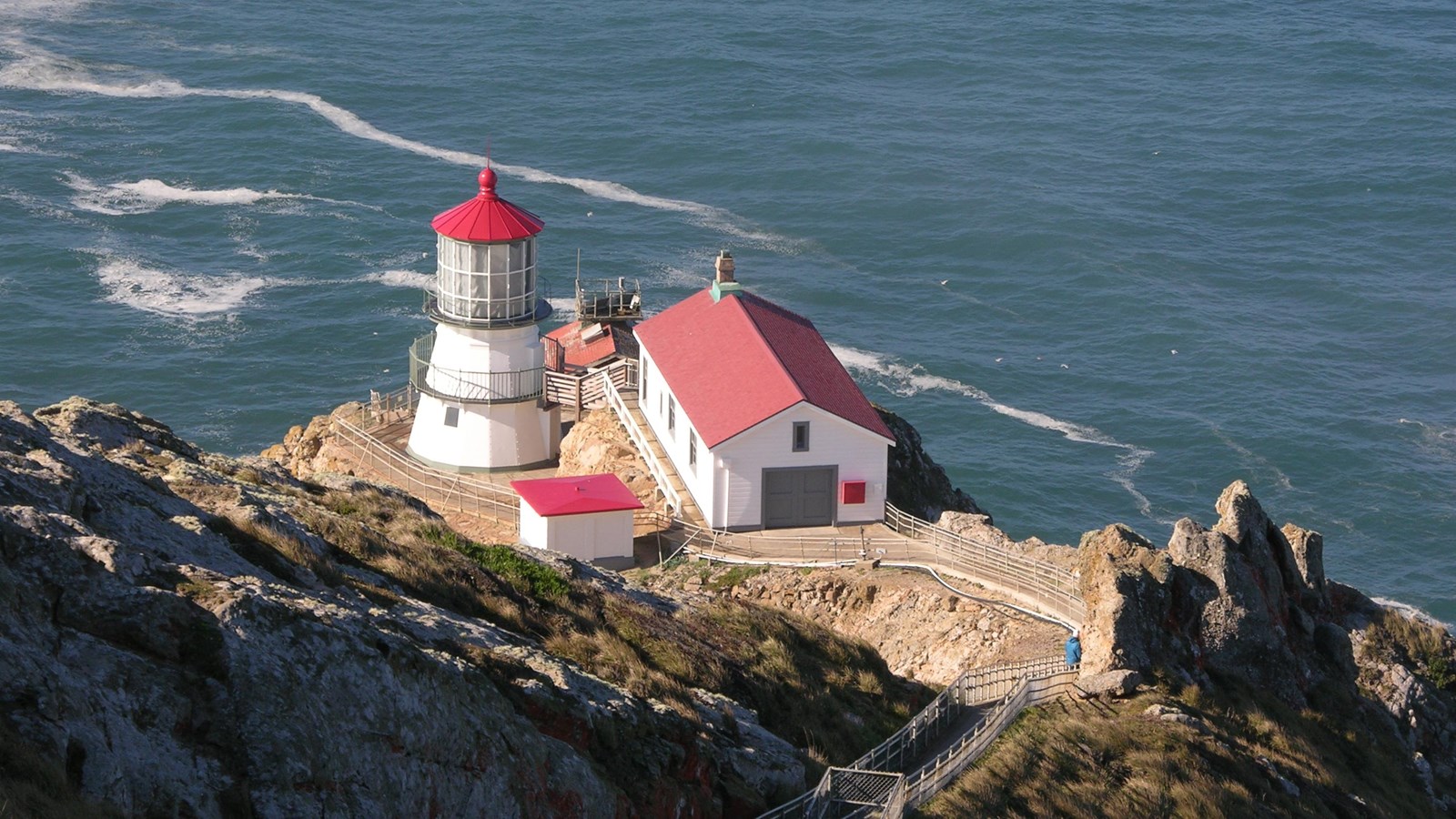 Three short, white-sided, red-roofed buildings sit on a rocky headland above the ocean.