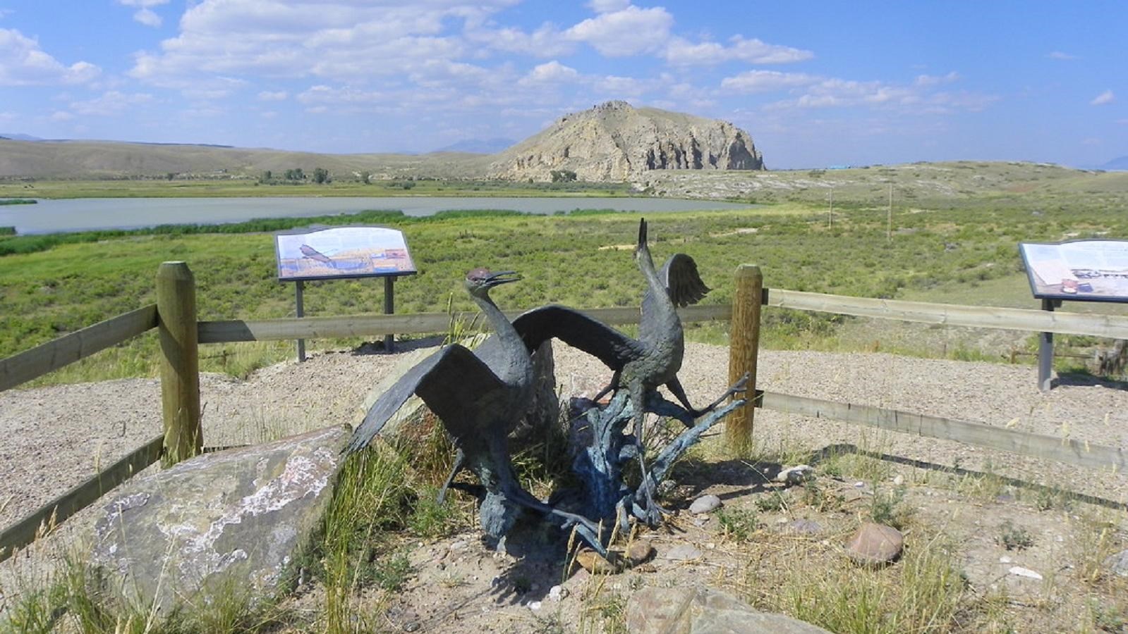 A statue of two cranes surrounded by a split rail fence and interpretive panels