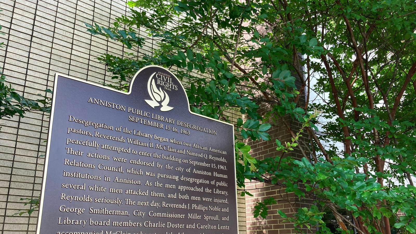 A brown and gold historic marker, near an overhanging tree. A grey stone wall fills the background.