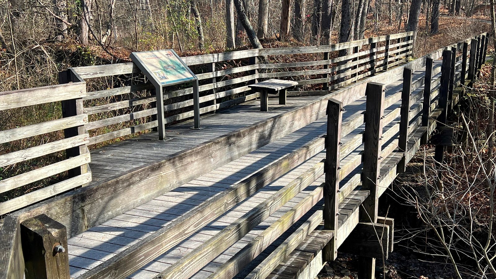 An angled view of a wooden bridge with a bench and exhibit panel, over a dry, woodland streambed.