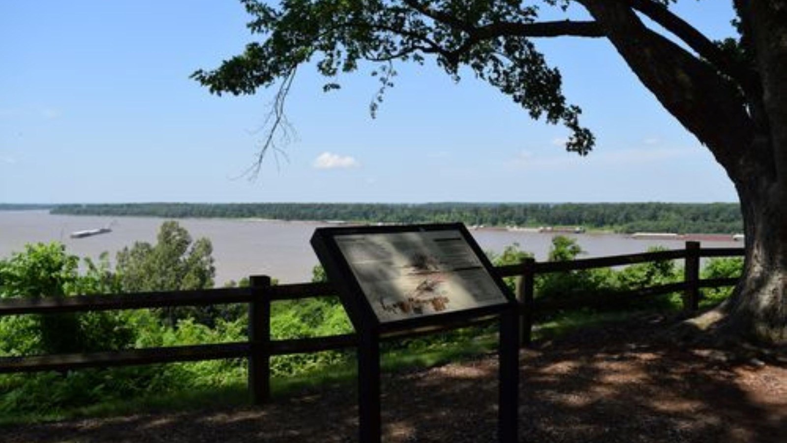 A sign sits next to a path overlooking a broad, flat river.