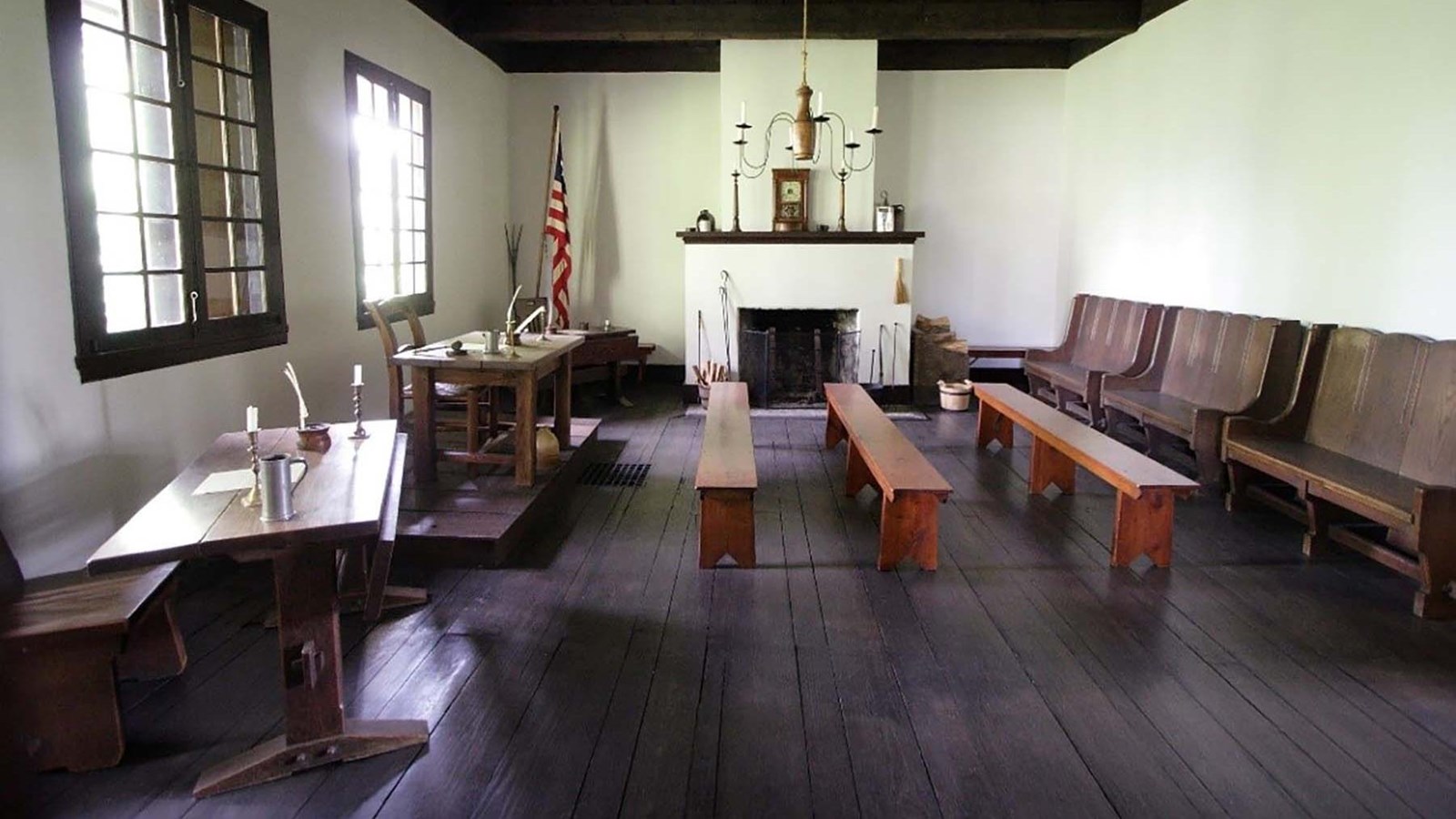 Large room with wooden floor, three benches in center of room facing two desks and american flag